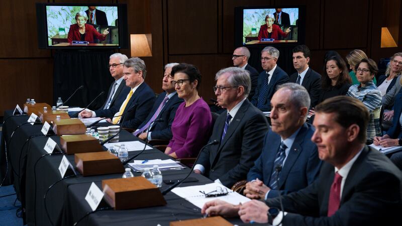 Sen. Elizabeth Warren, D-Mass., on a screen in the background, questions witnesses about...